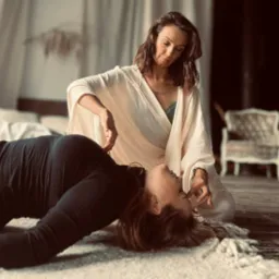 Kobieta w białym szlafroku delikatnie dotyka twarzy leżącej osoby, tworząc intymną atmosferę. Scena w jasnych, naturalnych barwach, z miękkim światłem.