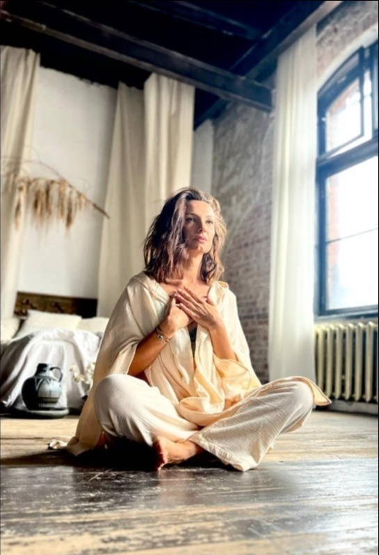 Kobieta w jasnym stroju siedzi po turecku w loftowym wnętrzu z ceglaną ścianą i dużym oknem, dłońmi dotyka klatki piersiowej. Obok łóżko i dzban.