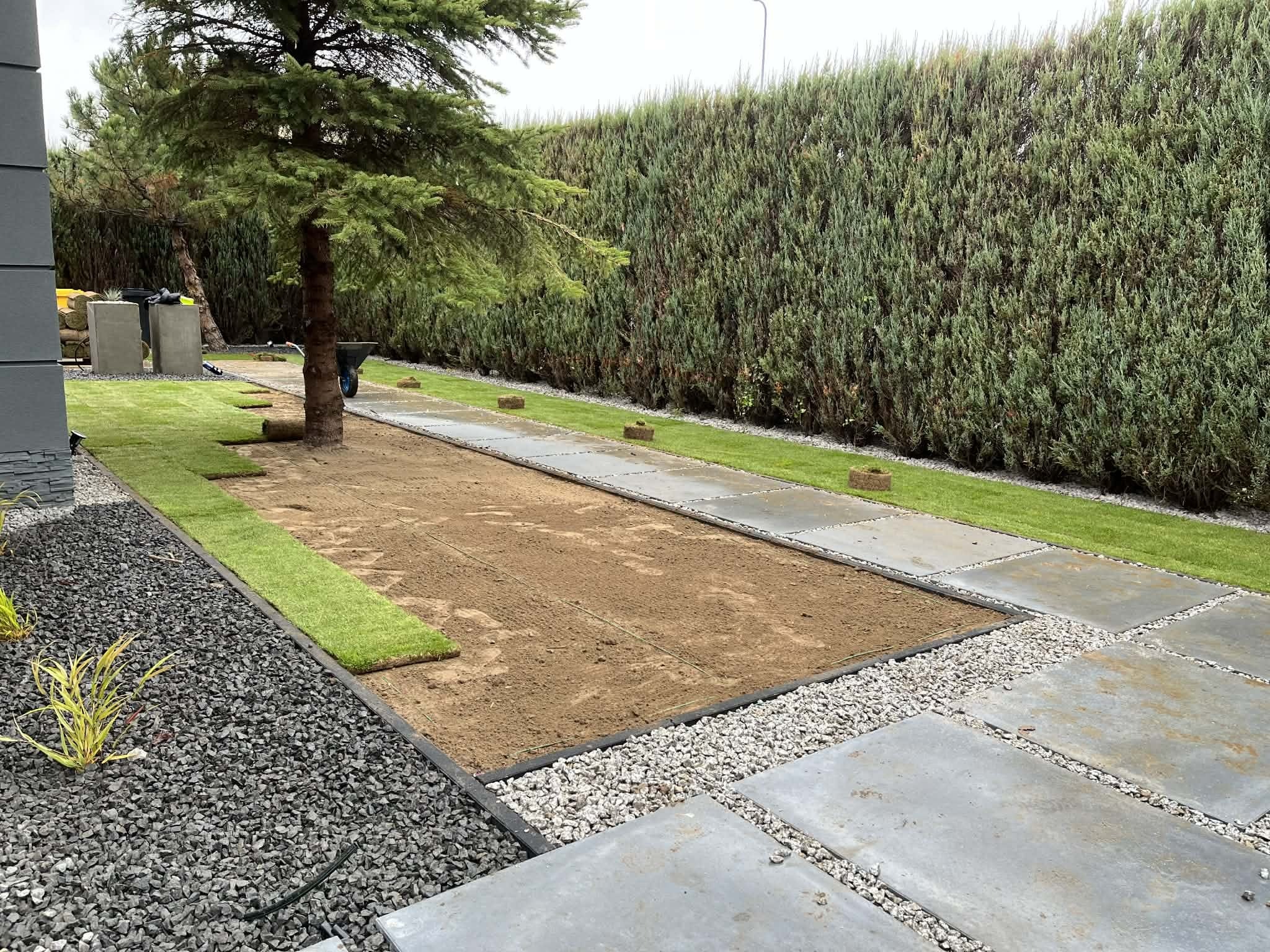 Układanie trawy z rolki w ogrodzie: widok na przygotowany teren, częściowo ułożone pasy trawy, obrzeża z kamienia i płyt, wysoki żywopłot w tle.