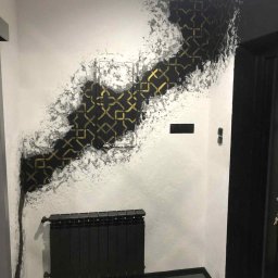 Art.Decor.Design - Wykończenie ściany z dekoracyjnym efektem ombre i geometrycznym wzorem w odcieniach czerni i złota, uzupełnione czarnym grzejnikiem w pomieszczeniu.