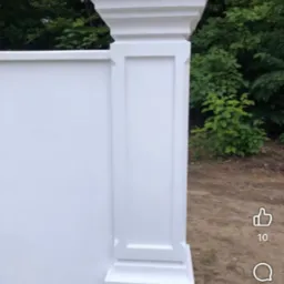 Biały, dekoracyjny filar z płyty karton-gips, imitujący kolumnę, na tle zieleni. Detal wykończenia z precyzyjnymi zdobieniami.