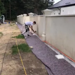 Pracownik w kombinezonie i masce maluje natryskowo fragment ogrodzenia posesji. Zabezpieczona folią i matą przestrzeń robocza. W tle dom z ciemnym dachem i drzewa.