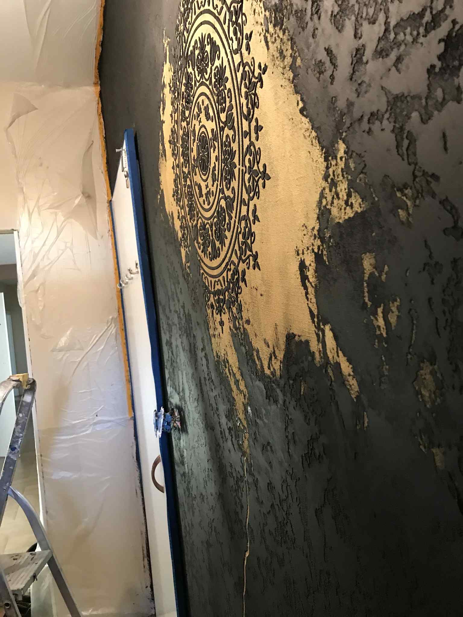 Dekoracyjna ściana w odcieniach szarości i złota z ornamentem, zabezpieczone drzwi i folia malarska, drabina. Efektowna aranżacja wnętrza w trakcie realizacji.