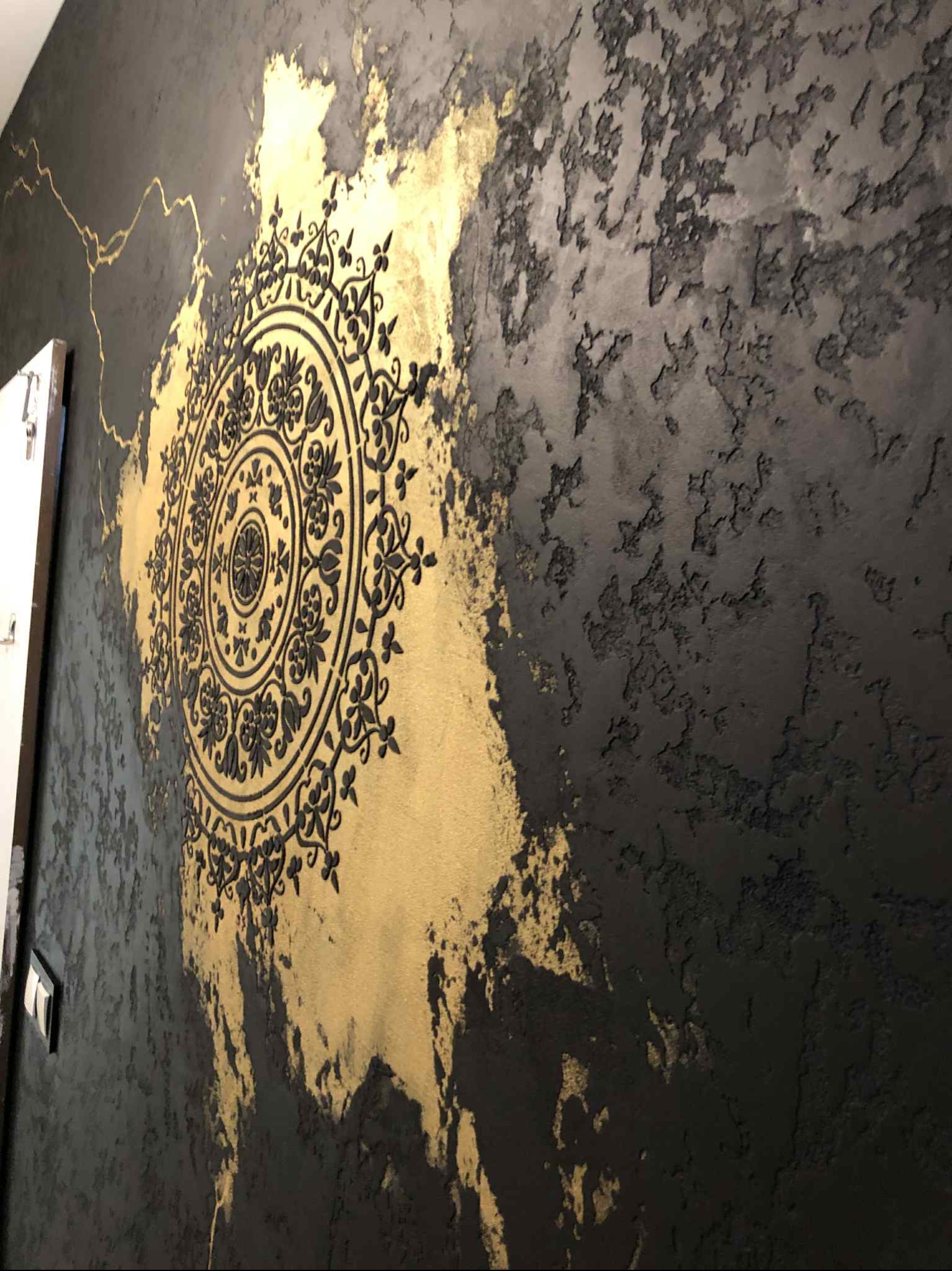Dekoracyjna ściana z efektem stiuku w odcieniach szarości i złota, zdobiona czarnym, wypukłym ornamentem w kształcie mandali. Widoczne detale faktury i nieregularne przejścia kolorystyczne.