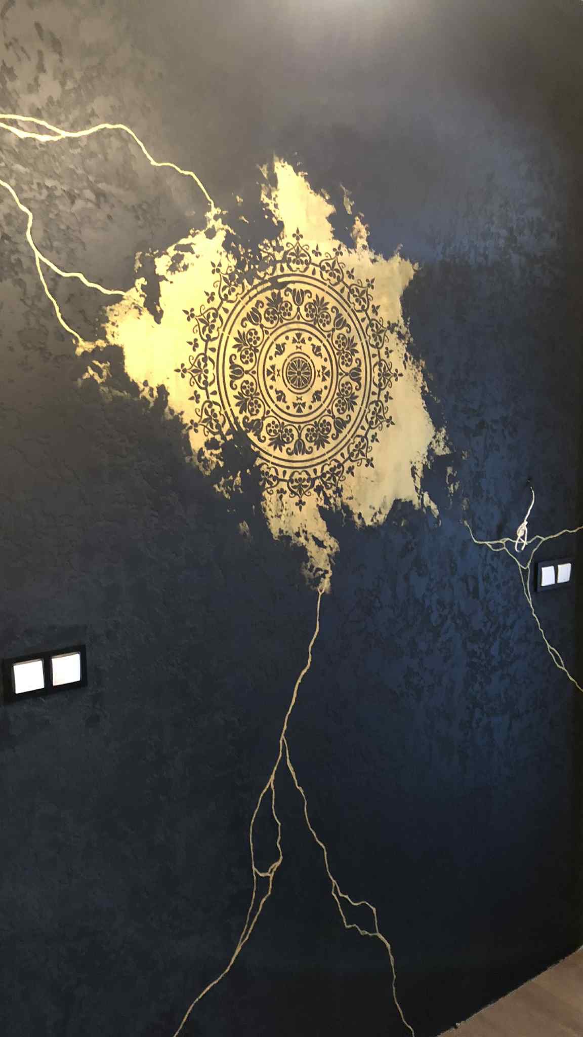 Dekoracyjna ściana w ciemnych barwach z efektem przetarć i złotym, okrągłym ornamentem oraz żyłkami, podświetlona przez dwa włączniki światła.