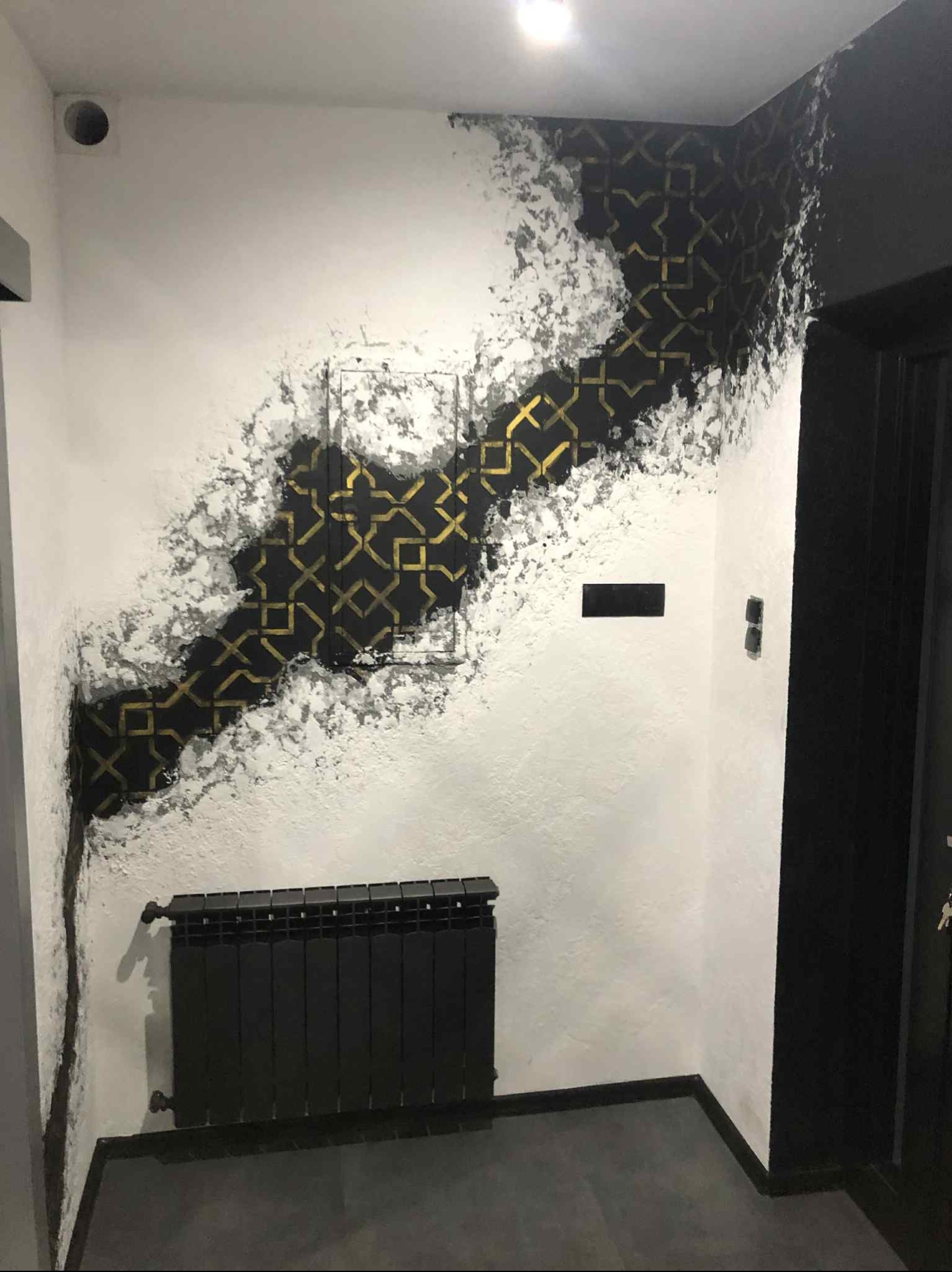 Pomysłowe malowanie ściany z motywem geometrycznym w kolorach czerni i złota, efektownie kontrastujące z bielą tła. Widoczny grzejnik w dolnej części kadru.