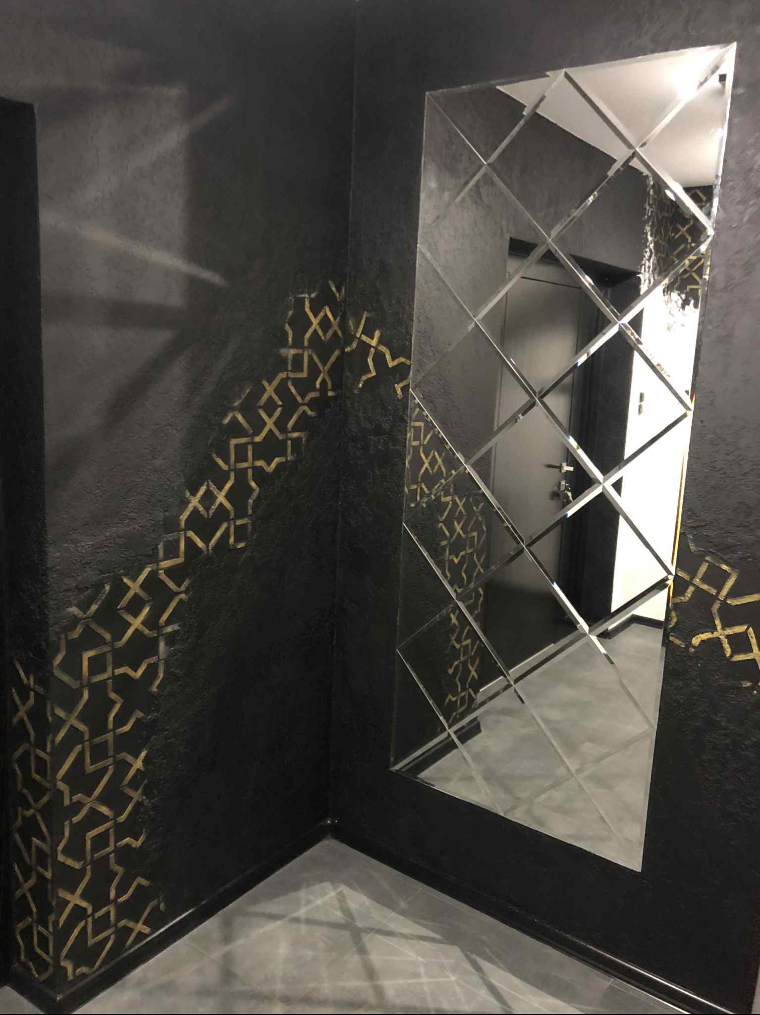 Ciemna ściana z dekoracyjnym wzorem w kolorze złota i lustro z geometrycznym szlifem, odbijające wnętrze pomieszczenia. Eleganckie wykończenie wnętrza.