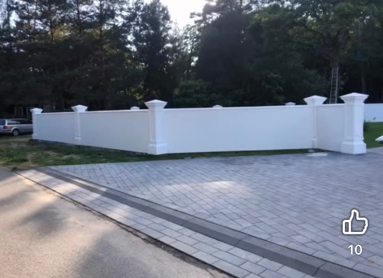 Biały, otynkowany mur z kolumnami dekoracyjnymi, widoczny z poziomu brukowanej nawierzchni. W tle drzewa i drabina oparta o mur.