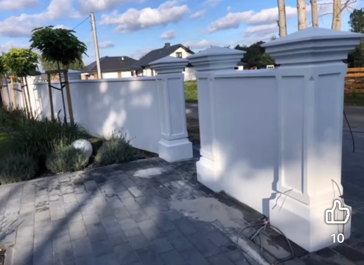 Biały, nowo pomalowany mur z dekoracyjnymi słupkami na tle zieleni i nieba. Świeżo ułożona kostka brukowa, widoczne ślady wody po myciu. Słupki z dekoracyjnymi kapitelami.