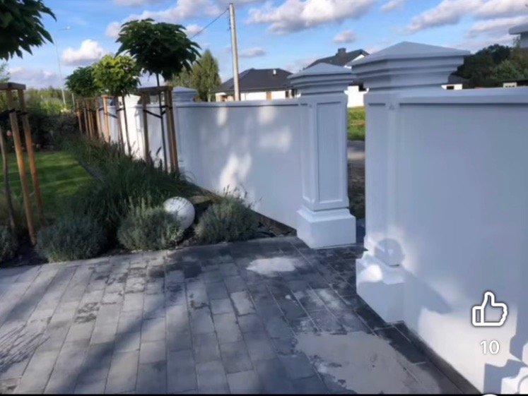 Biały, dekoracyjny mur z kolumnami i nasadami w ogrodzie, obok ścieżka z szarej kostki brukowej, drzewa wzdłuż muru. Eleganckie wykończenie.