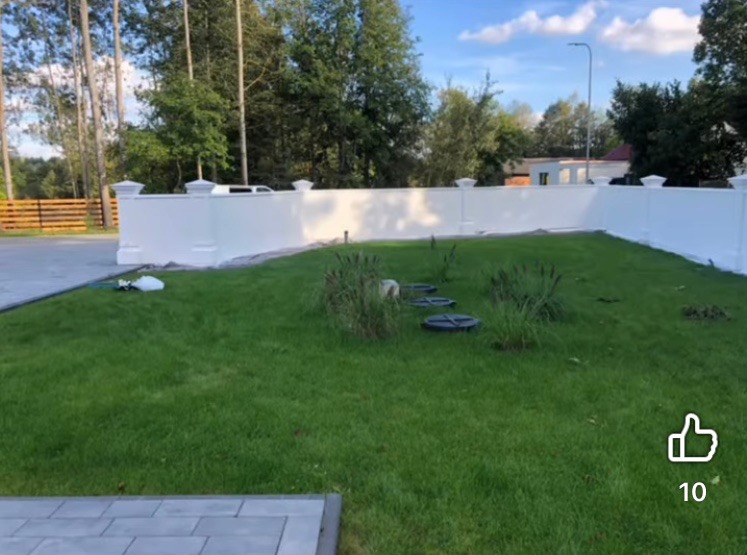 Biały, pomalowany mur z dekoracyjnymi słupkami na tle zielonego trawnika i drzew. Widoczne studzienki kanalizacyjne i fragment podjazdu z kostki.