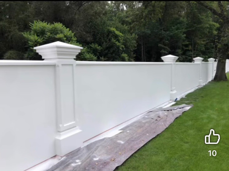Biały, pomalowany mur z dekoracyjnymi słupkami na tle zieleni. Widoczne zabezpieczenia malarskie u dołu muru.
