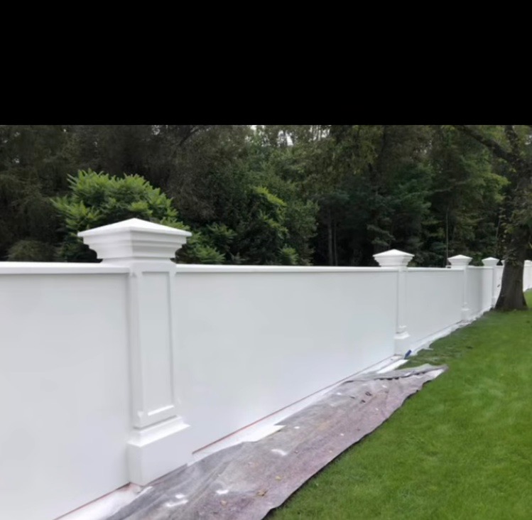 Biały, dekoracyjny mur ogrodzeniowy z kolumnami i zadaszeniem, świeżo pomalowany, z widocznym zabezpieczeniem na trawie. Otoczenie: zieleń drzew i trawnik.