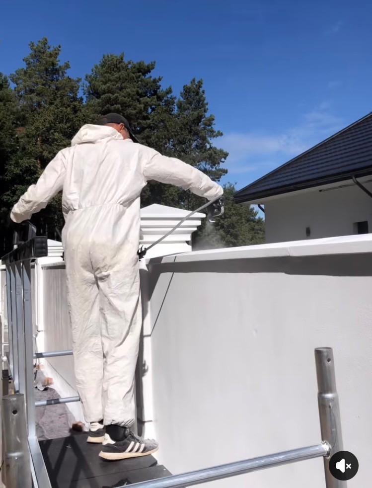 Malowanie natryskowe ogrodzenia w Złoczewie. Pracownik w kombinezonie maluje białą farbą. Widoczne rusztowanie i fragment budynku. Błękitne niebo w tle.