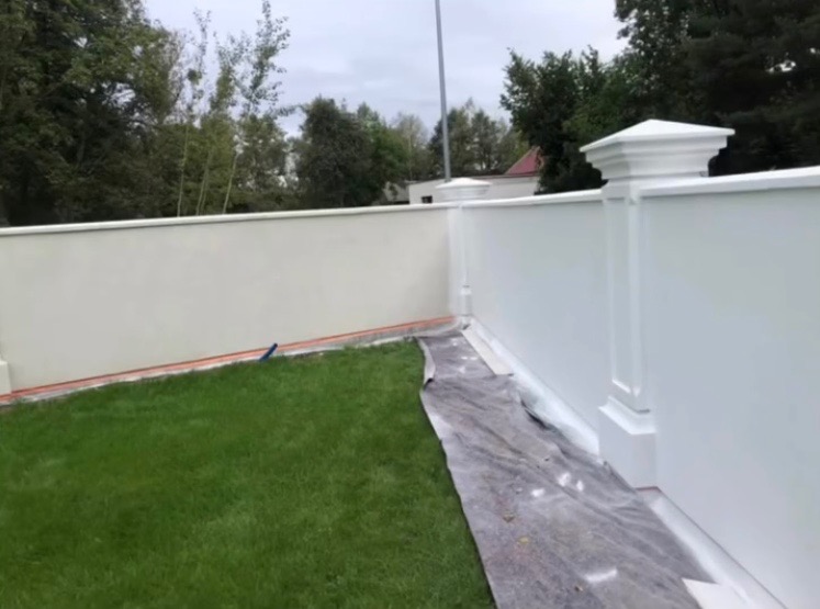 Świeżo pomalowany, biały mur z dekoracyjnymi kolumnami w ogrodzie. Część muru w odcieniu kremowym, zabezpieczona folią malarską. Soczysta zieleń trawnika.