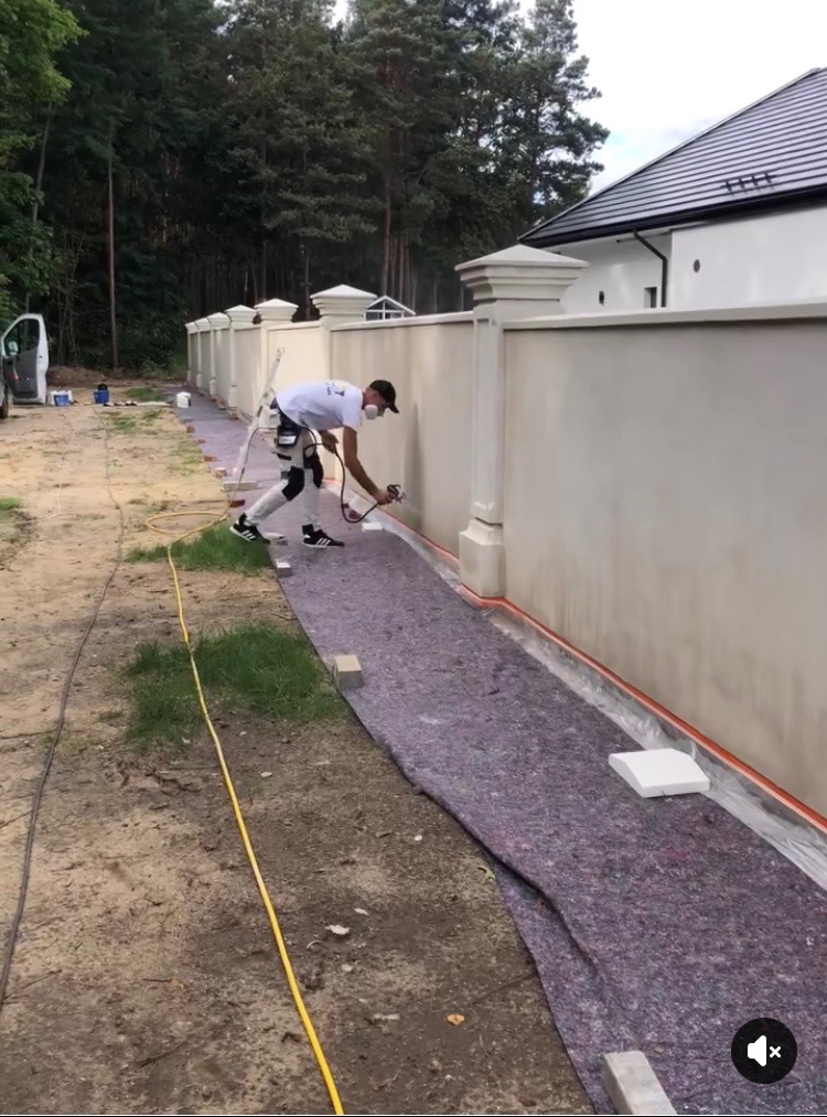 Pracownik w kombinezonie i masce maluje natryskowo fragment ogrodzenia posesji. Zabezpieczona folią i matą przestrzeń robocza. W tle dom z ciemnym dachem i drzewa.