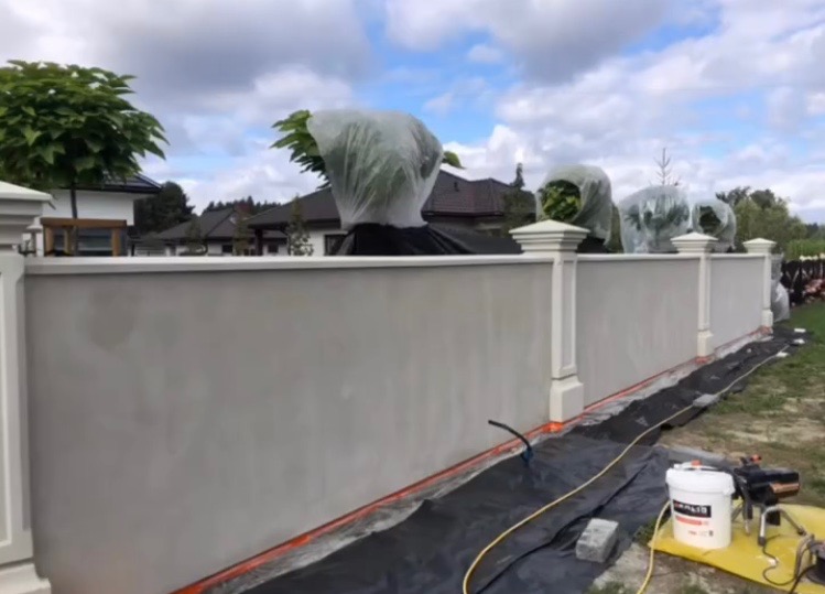 Świeżo otynkowany, jasnoszary mur ogrodzeniowy z ozdobnymi słupkami. W tle zabudowa jednorodzinna i drzewa. Na ziemi folia ochronna i sprzęt malarski.