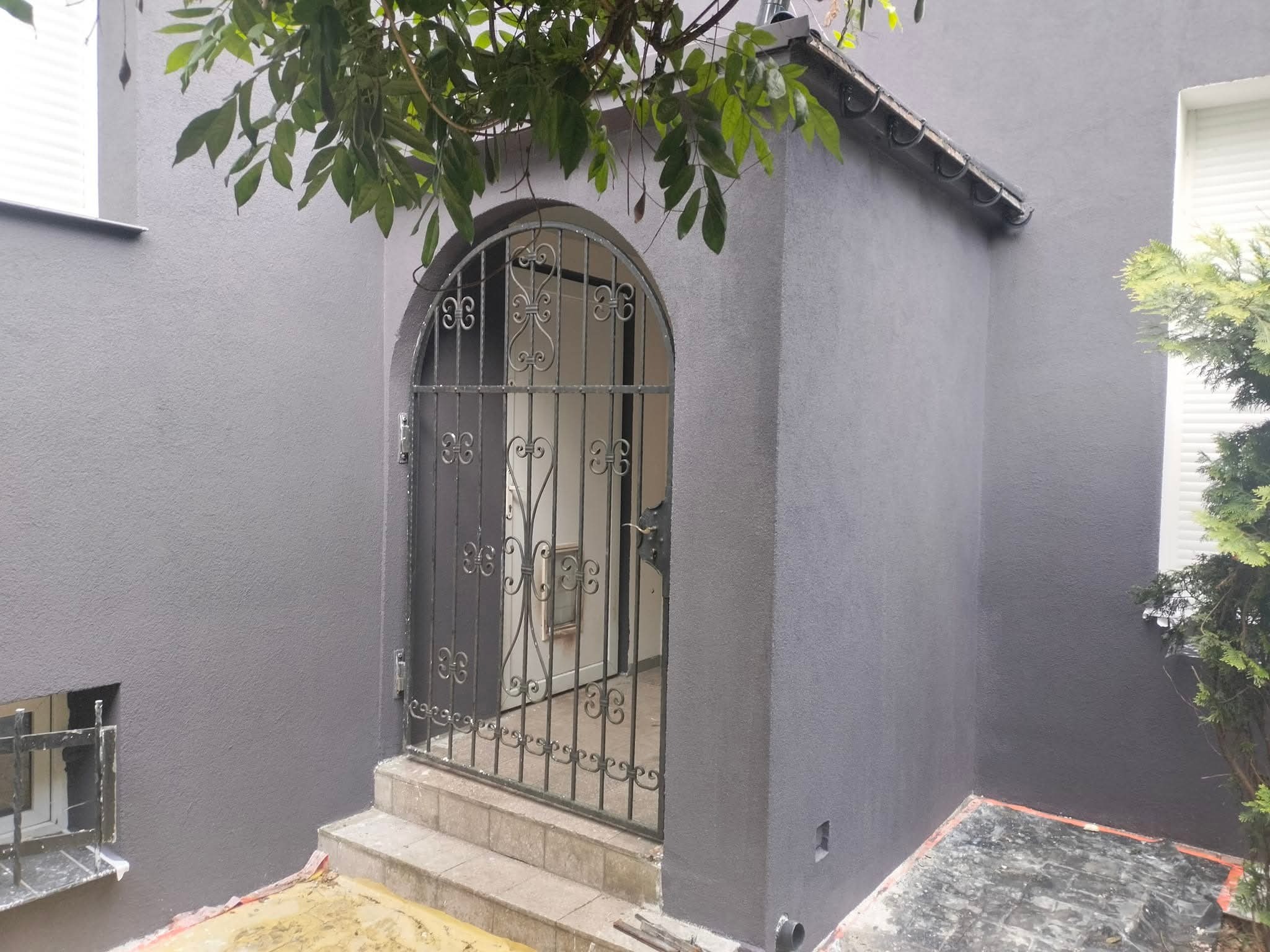Szara elewacja domu z ozdobną, metalową bramką wejściową. Widoczne świeżo pomalowane ściany i fragment roślinności nad bramką.