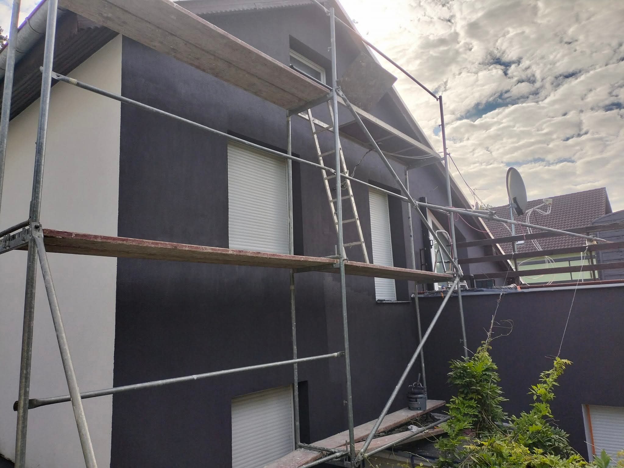Malowanie elewacji domu na ciemny kolor. Widoczne rusztowania i fragment pomalowanej na biało ściany. Częściowo zasłonięte okna z roletami.