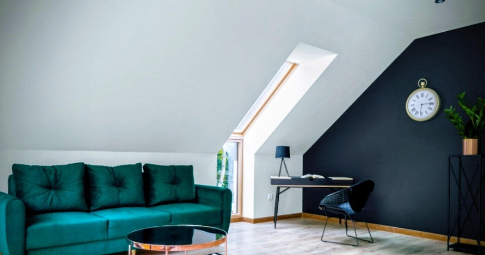Stylowe poddasze z oknem dachowym: szmaragdowa sofa, czarne biurko, zegar na granatowej ścianie. Jasne panele podłogowe i roślina w złotej doniczce.