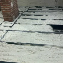 Poddasze po izolacji pianką: widoczne krokwie i mur z cegły, pokryte grubą warstwą białej pianki. Detal: nierównomierna struktura piany.