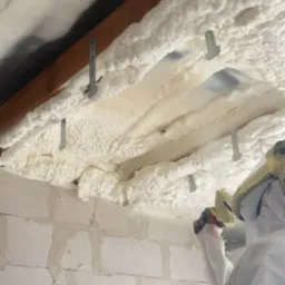 Prace izolacyjne pianą PUR na poddaszu: biała piana aplikowana na drewniane belki, metalowe mocowania, fragment ściany z bloczków i osoba w kombinezonie ochronnym.