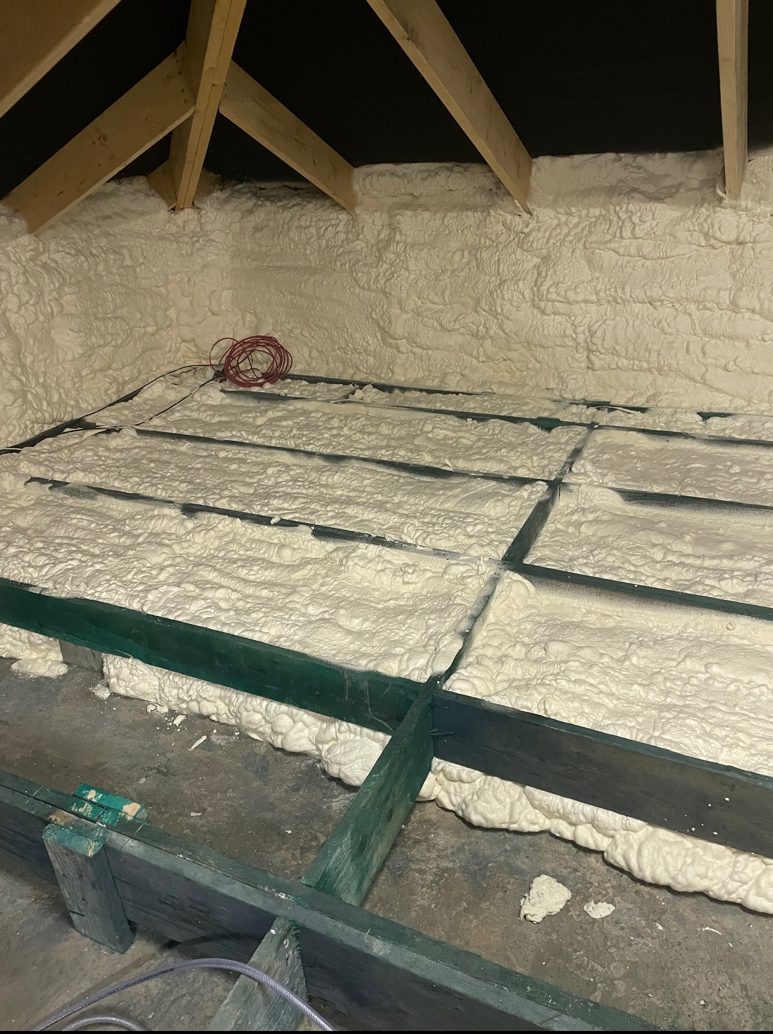 Poddasze ocieplone pianką PUR, widoczne drewniane krokwie i zielona konstrukcja podłogi. Na podłodze leży zwój czerwonego kabla.