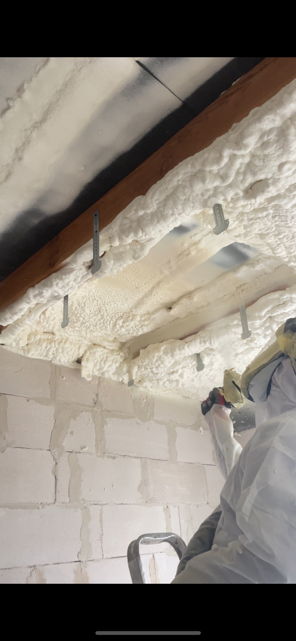 Prace izolacyjne pianą PUR na poddaszu: biała piana aplikowana na drewniane belki, metalowe mocowania, fragment ściany z bloczków i osoba w kombinezonie ochronnym.