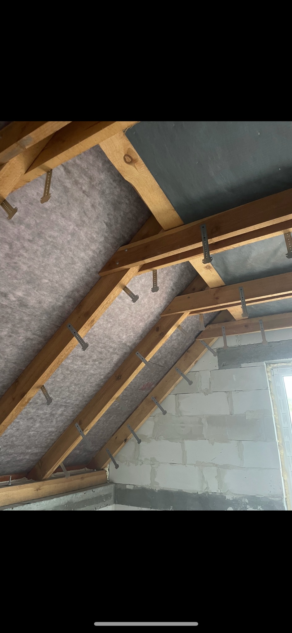 Poddasze z drewnianą konstrukcją, izolacją i metalowymi uchwytami. Ściany z białych bloczków, fragment okna. Prace izolacyjne w trakcie realizacji.