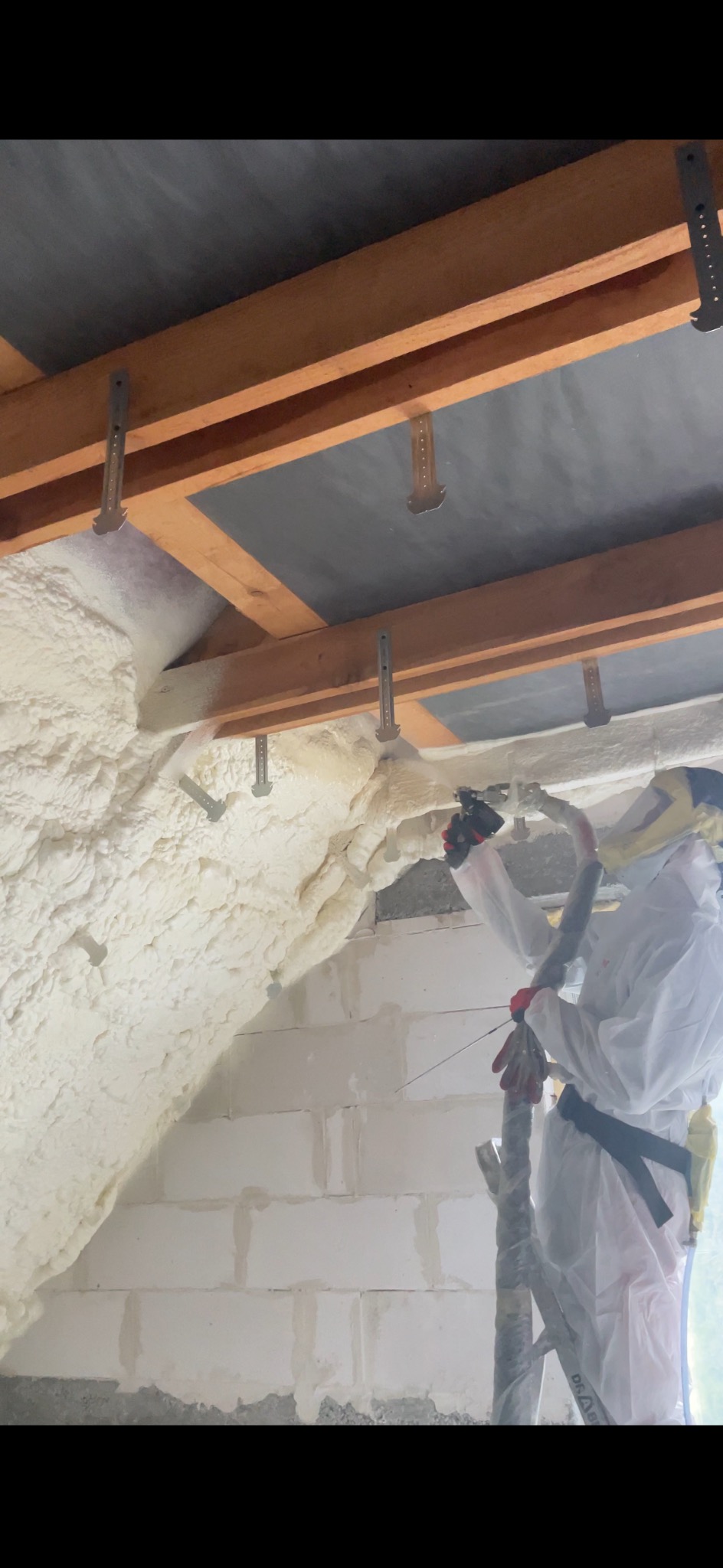 Pracownik w kombinezonie nanosi pianę izolacyjną na poddasze, widoczne drewniane elementy konstrukcyjne i metalowe mocowania. Świeża, jasna piana pokrywa skośną powierzchnię.