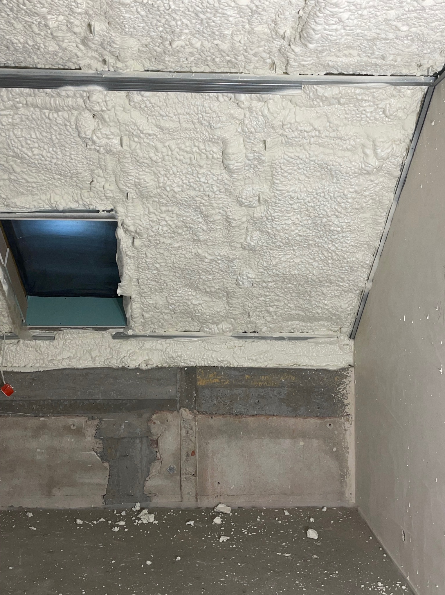 Poddasze po natrysku pianki PUR, widoczne metalowe profile i fragment okna dachowego. Ściany częściowo przygotowane do wykończenia, surowy beton na dole.