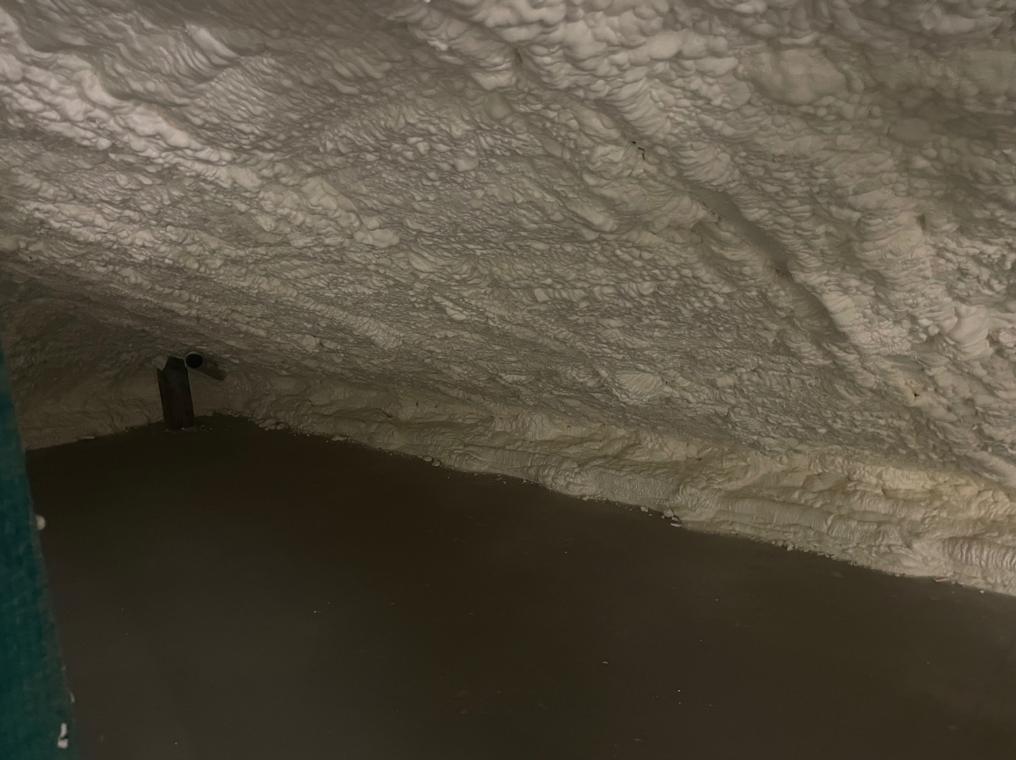 Poddasze po ociepleniu pianką PUR, widoczne nierównomierne nałożenie izolacji, ciemna podłoga i fragment instalacji. Charakterystyczna struktura piany.