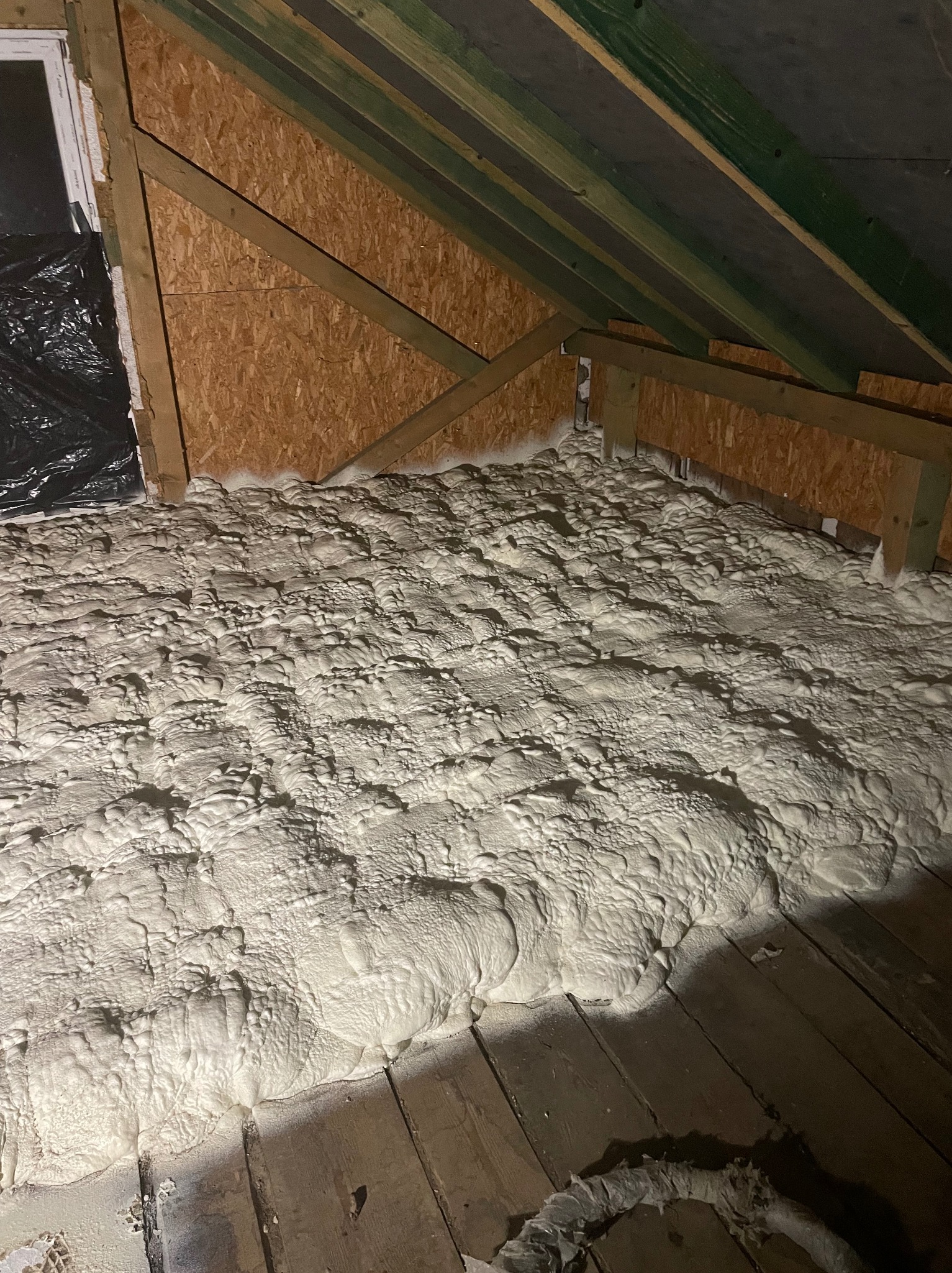 Poddasze z drewnianą konstrukcją i deskami, wypełnione białą pianą izolacyjną, szczelnie pokrywającą powierzchnię. Widoczne fragmenty okna i czarnej folii.