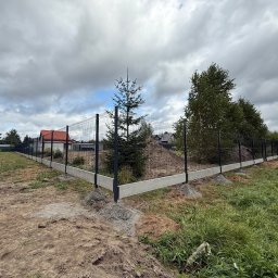 Kermit Ogrodzenia Piotr Sałański - Czarny panel ogrodzeniowy z podmurówką betonową, widok z narożnika działki. W tle dom z czerwoną dachówką i pochmurne niebo. Na pierwszym planie ziemia i trawa.