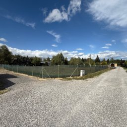 Kermit Ogrodzenia Piotr Sałański - Siatka ogrodzeniowa otacza działkę z utwardzoną drogą, błękitne niebo z chmurami w tle. Widoczne drzewa i budynki w oddali. Perspektywa z poziomu gruntu.