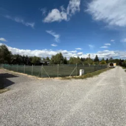 Siatka ogrodzeniowa otacza działkę z utwardzoną drogą, błękitne niebo z chmurami w tle. Widoczne drzewa i budynki w oddali. Perspektywa z poziomu gruntu.