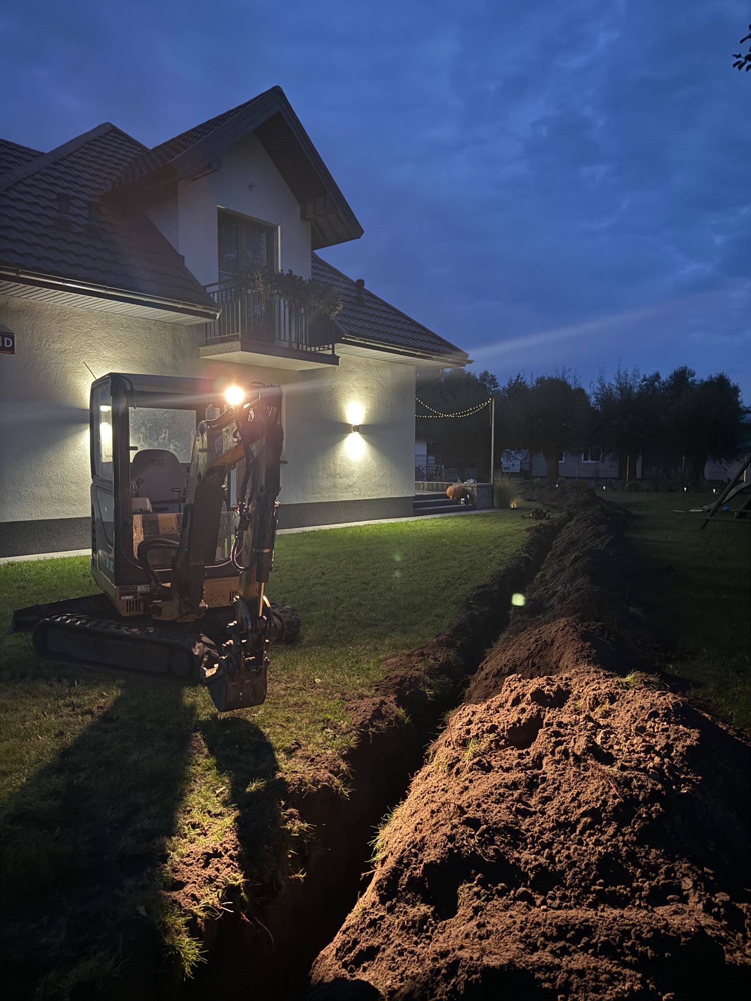 Mała koparka przy wykopie w ziemi, oświetlona światłami, na tle domu z balkonem i dekoracyjnymi lampkami w ogrodzie, nocne ujęcie.
