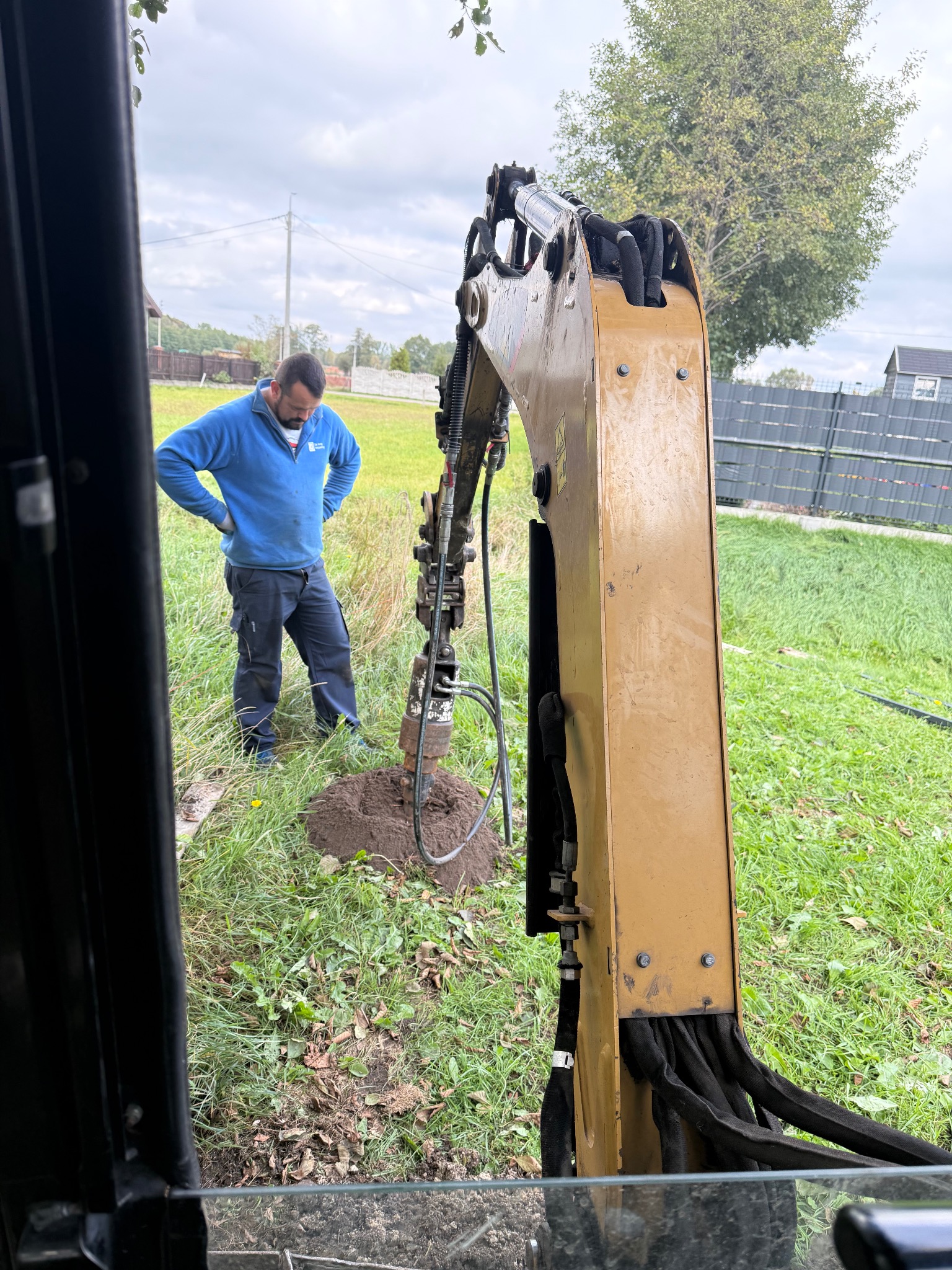 Wykop pod fundamenty w Woli Cygowskiej: Operator mini-koparki, w niebieskiej bluzie, nadzoruje wiercenie otworu w ziemi pod ogrodzenie na zielonej działce.