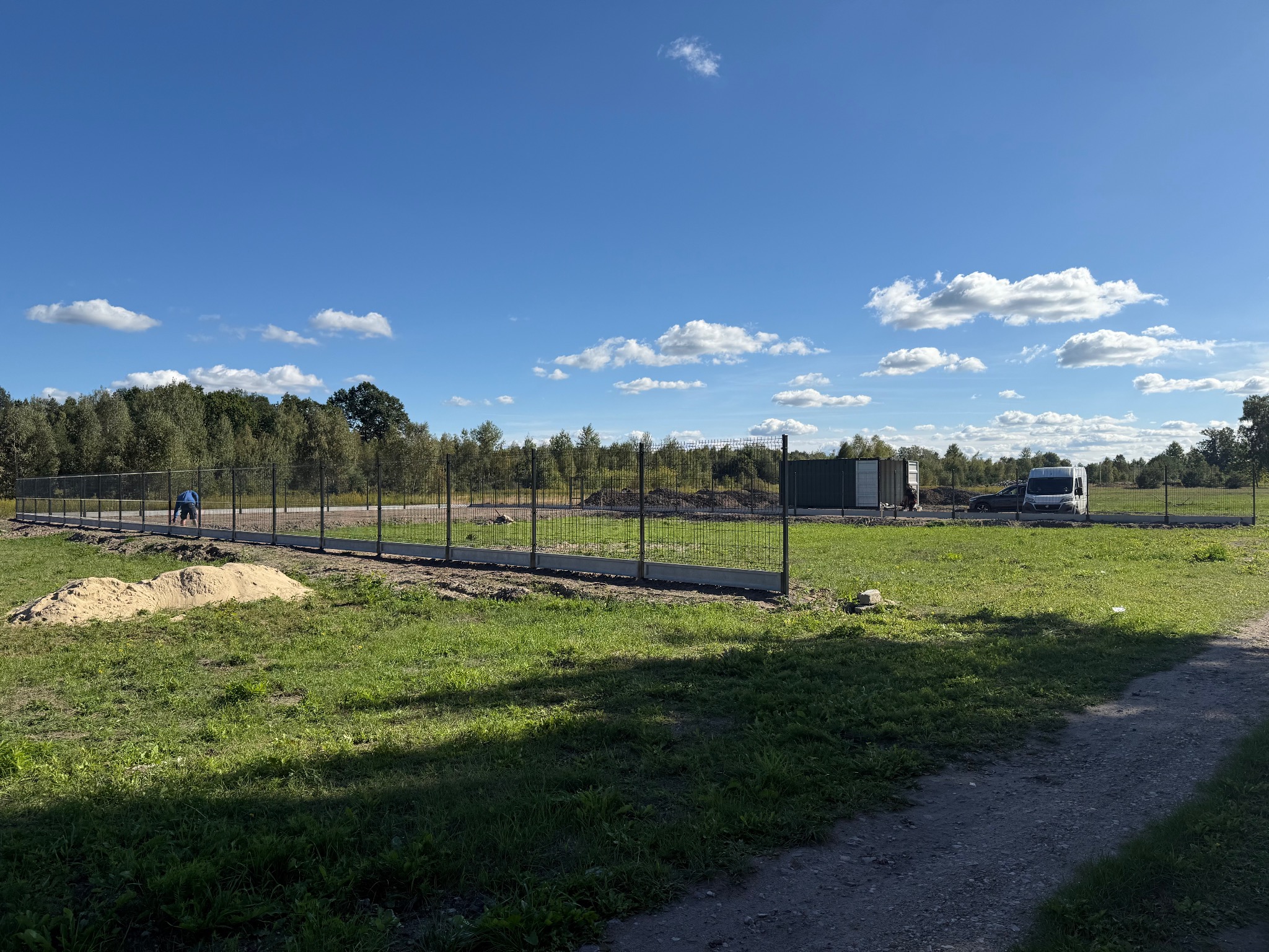 Montaż panelowego ogrodzenia na fundamencie, widok na teren budowy z pracownikiem w oddali, błękitne niebo z chmurami w tle.