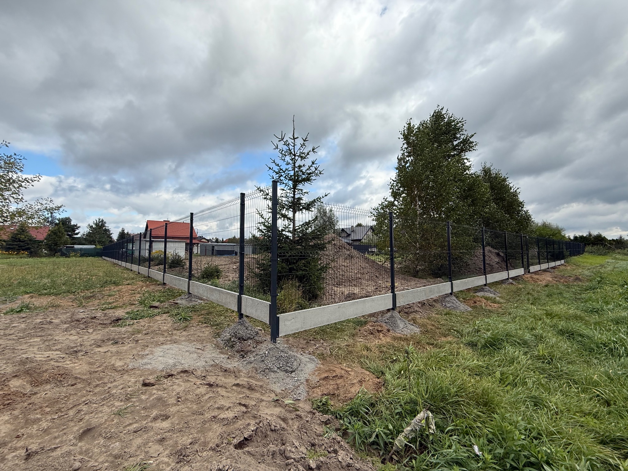 Czarny panel ogrodzeniowy z podmurówką betonową, widok z narożnika działki. W tle dom z czerwoną dachówką i pochmurne niebo. Na pierwszym planie ziemia i trawa.