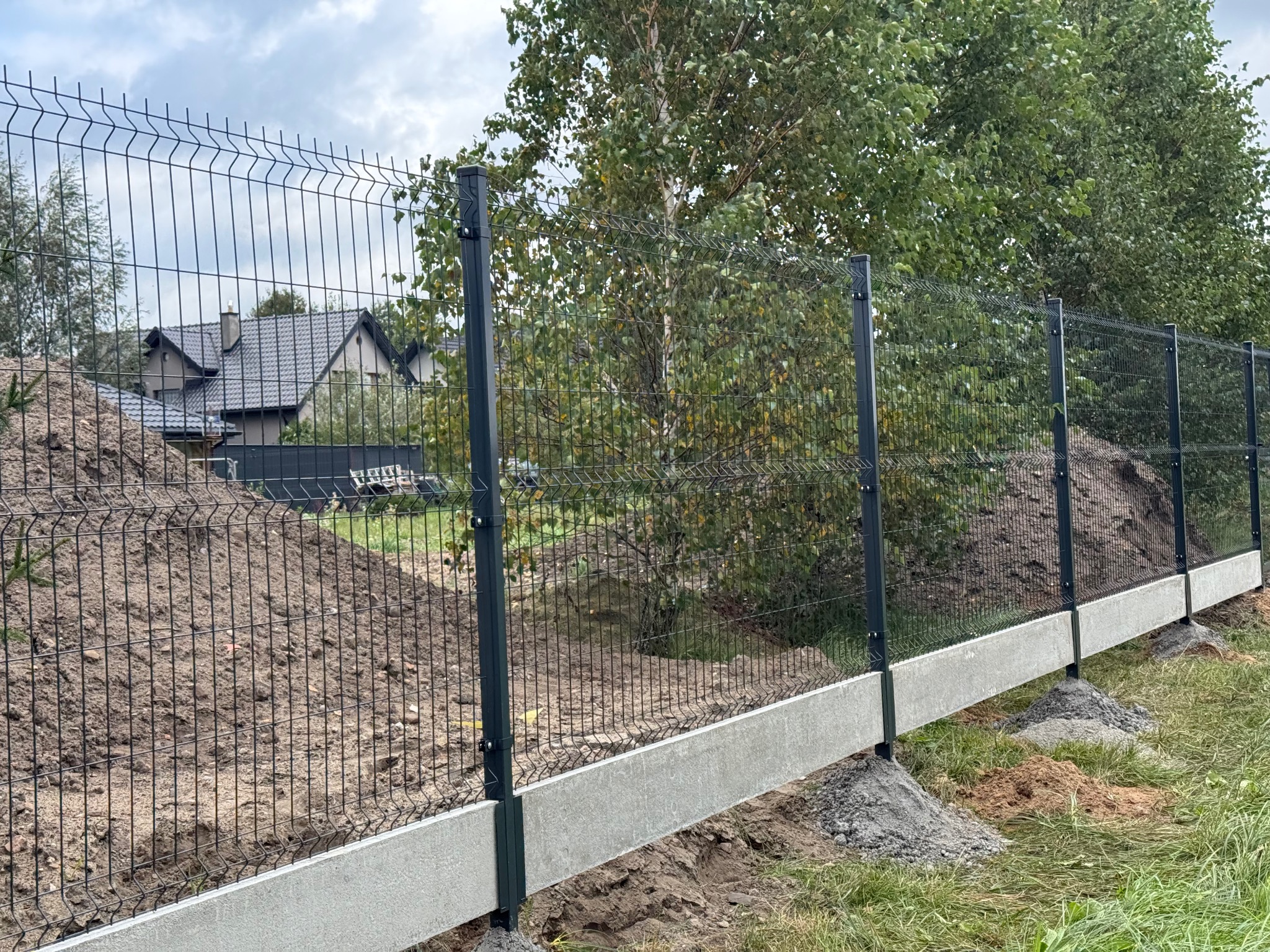 Nowo zamontowane ogrodzenie z siatki panelowej na betonowej podmurówce, widoczne słupki i fragment domu w tle, na trawie pozostałości po pracach ziemnych.