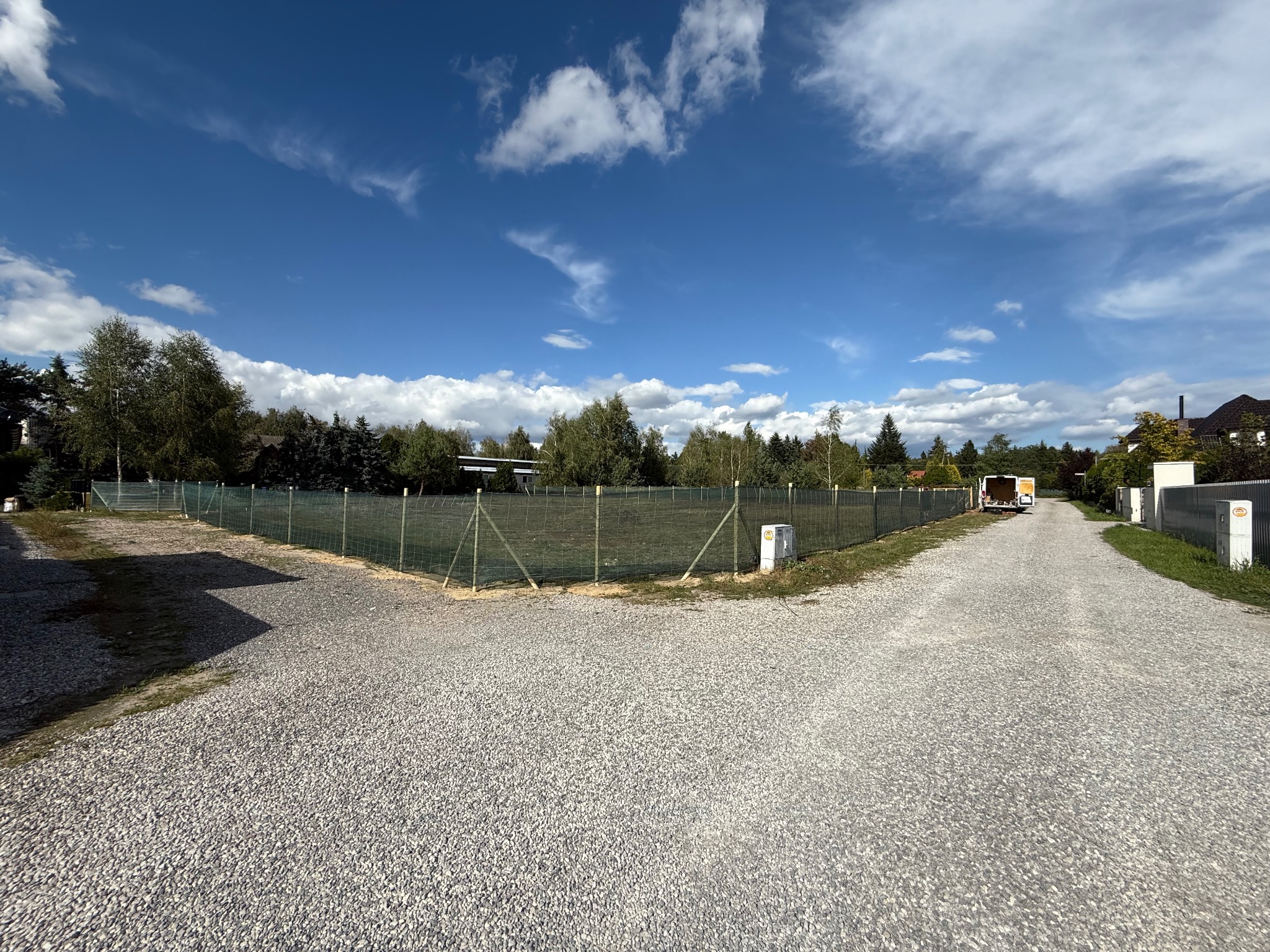 Siatka ogrodzeniowa otacza działkę z utwardzoną drogą, błękitne niebo z chmurami w tle. Widoczne drzewa i budynki w oddali. Perspektywa z poziomu gruntu.