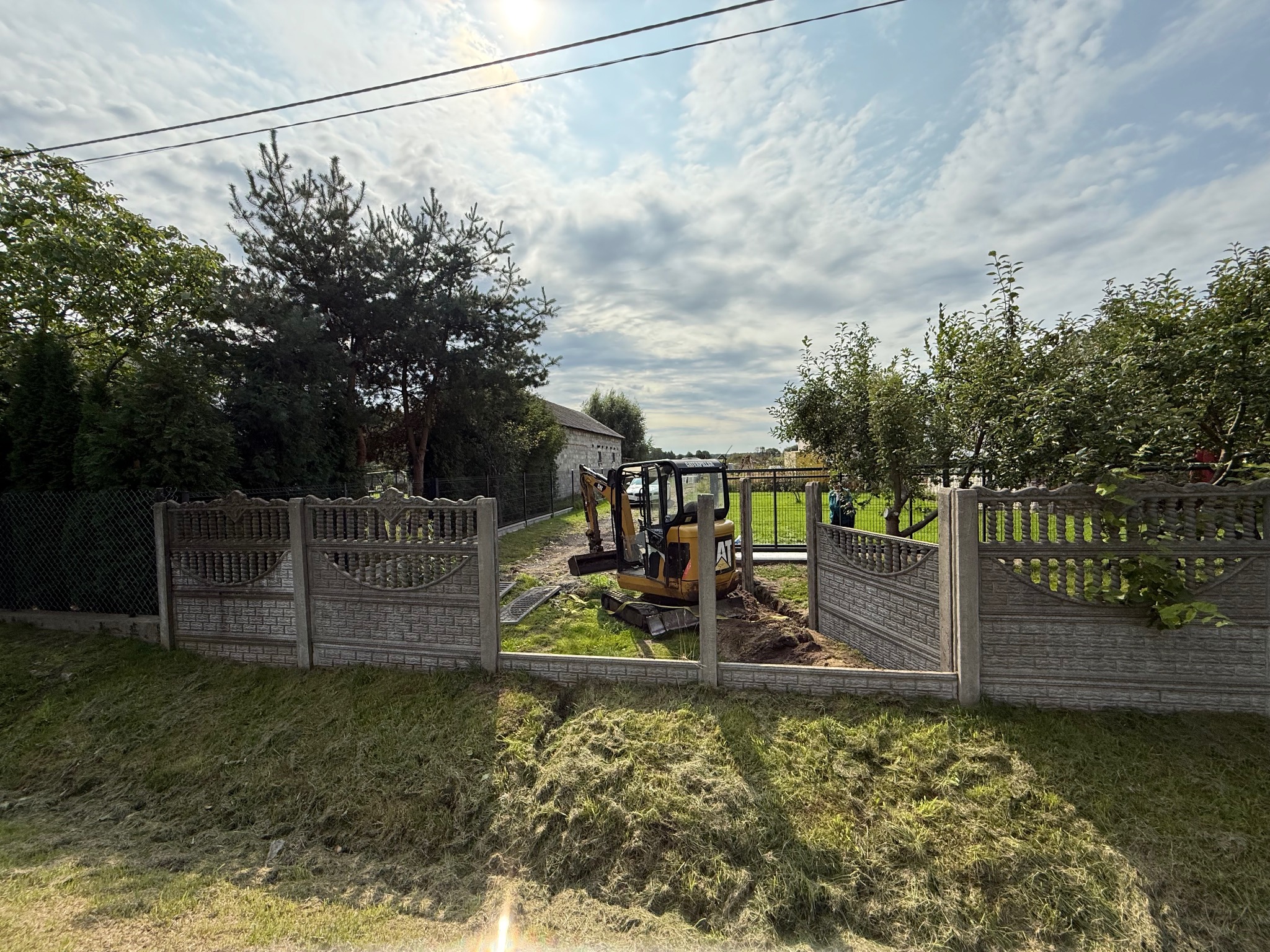 Mini koparka CAT na trawniku, częściowo rozkopana ziemia między przęsłami betonowego ogrodzenia, w tle budynek gospodarczy, błękitne niebo z chmurami.