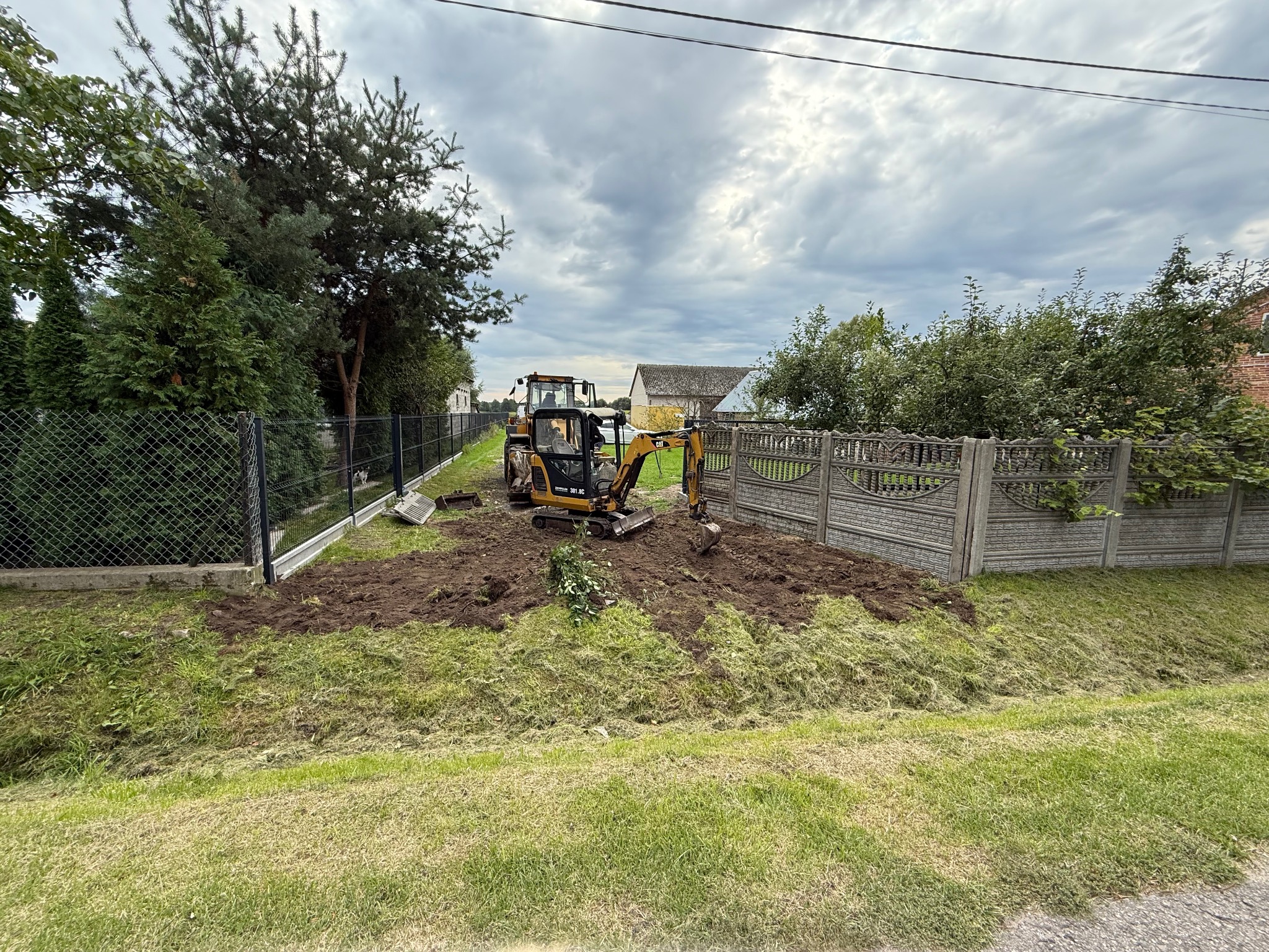 Żółta koparka CAT na działce, w tle widoczny traktor i ogrodzenia panelowe oraz siatka. Przygotowanie terenu pod nasadzenia, wykop ziemi.