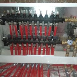 Dmytro Zlenko - Rozdzielacz ogrzewania podłogowego w metalowej szafce, z czerwonymi rurami, manometrami i pompą obiegową. Widoczne datowniki na podłodze.