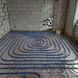 Dmytro Zlenko - Niebieskie rury ogrzewania podłogowego ułożone spiralnie na czerwonej siatce w surowym wnętrzu z białego bloczka, widok z góry, etap instalacji przed wylewką.