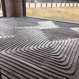 Dmytro Zlenko - Ułożona instalacja ogrzewania podłogowego w pokoju z dużym oknem i ceglanym filarem. Widoczne rury grzewcze na folii izolacyjnej przed wylaniem posadzki.