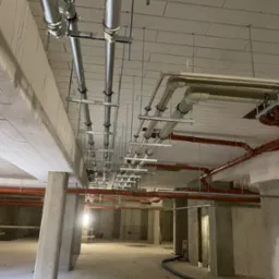 Widok instalacji pod stropem: rury stalowe i PCV, podwieszone na metalowych konstrukcjach. Stan surowy, brak wykończenia wnętrz. Widoczna perspektywa długiego korytarza.