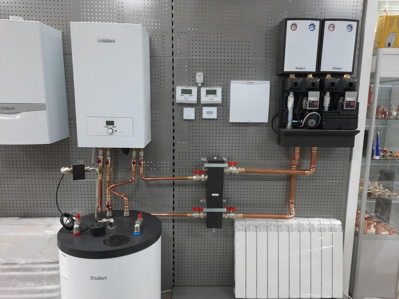 Kompletna instalacja grzewcza Vaillant: kotły, zbiornik, grzejnik. Miedziane rury łączą elementy na tle perforowanej ściany ekspozycyjnej.