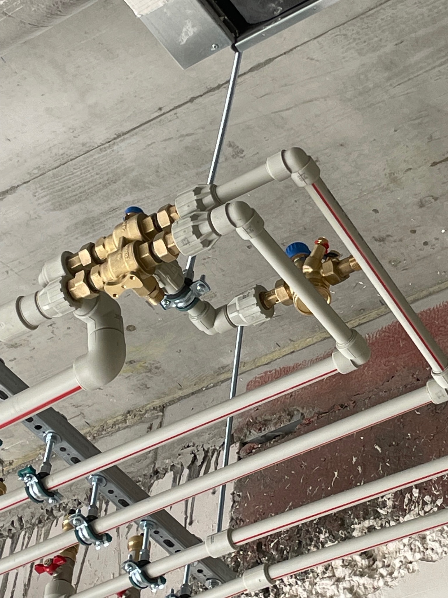 Szare i białe rury instalacji wodnej z mosiężnymi zaworami, zamontowane pod betonowym sufitem, widoczne mocowania i elementy instalacji.